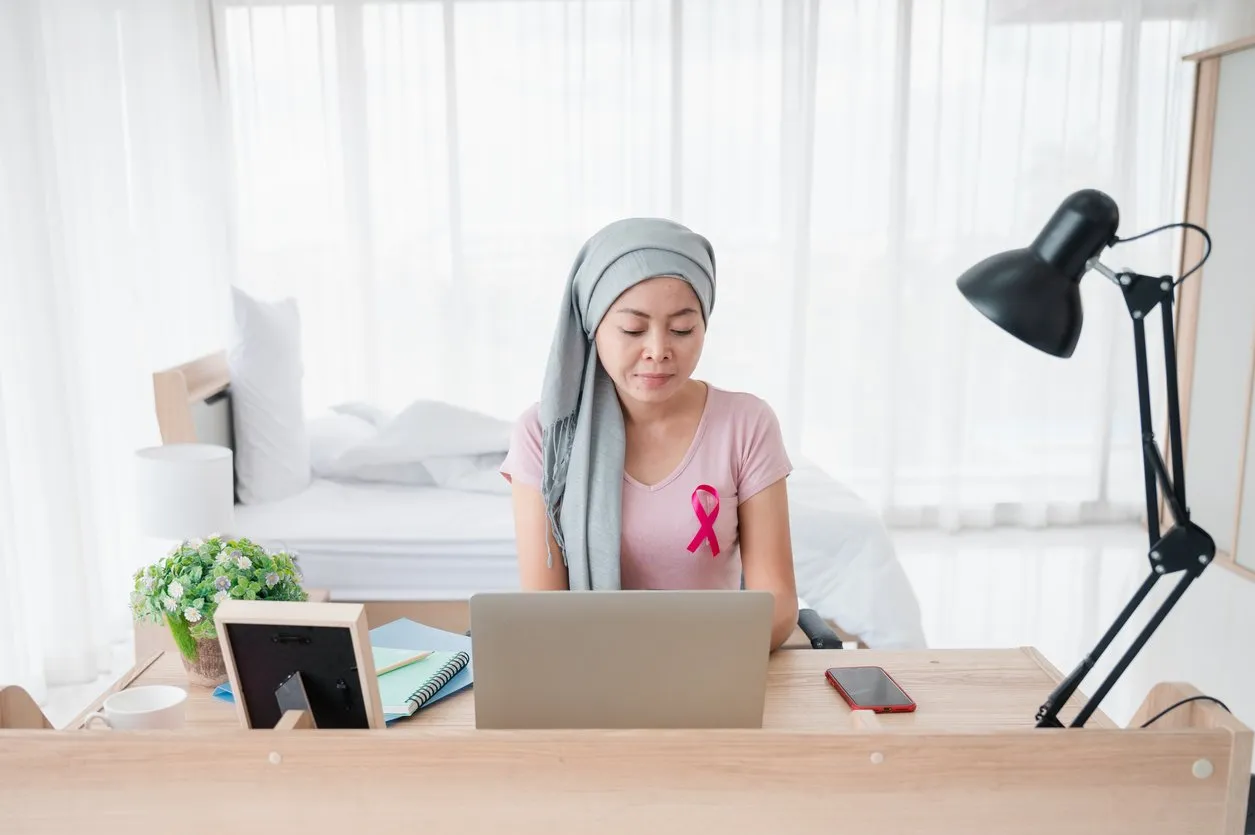 woman wearing a hijab uses smartphone and laptop at her desk. Attaching a pink ribbon represents recovery from a breast cancer patient