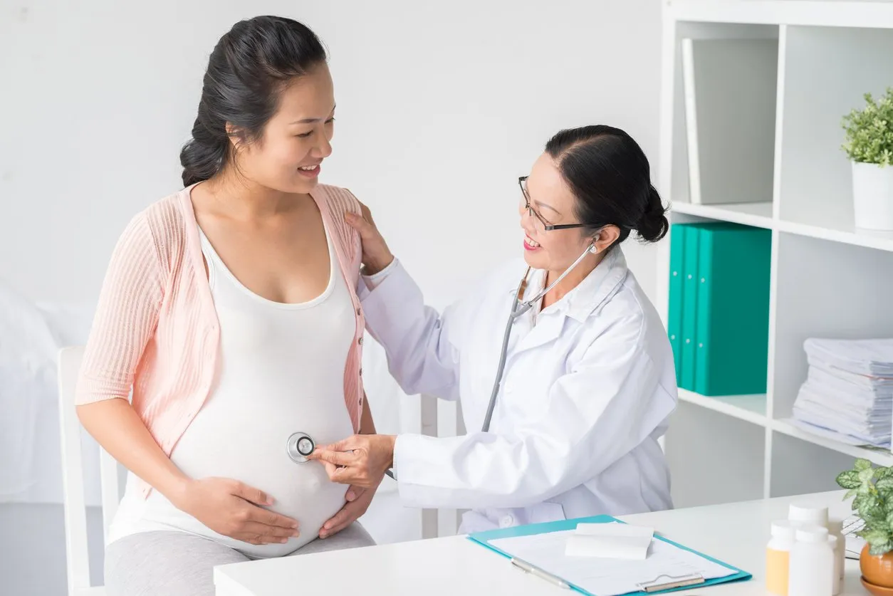 Asian doctor with stethoscope listening to belly of pregnant woman